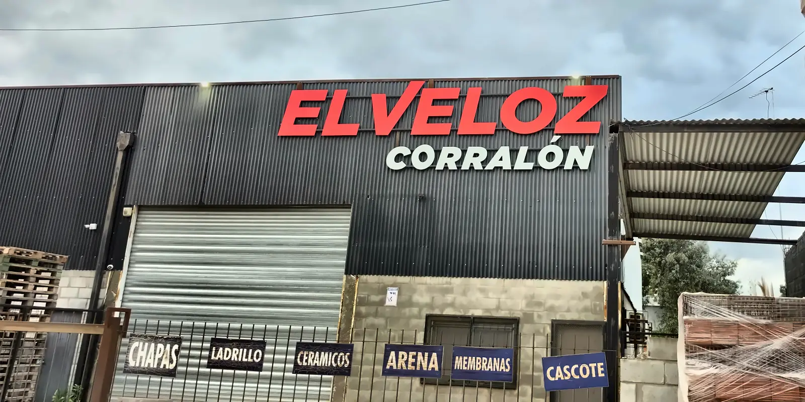 Frente de Corralón El Veloz con el cartel y los carteles de rubros: chapas, ladrillo, cerámicos, arena, membranas y cascote